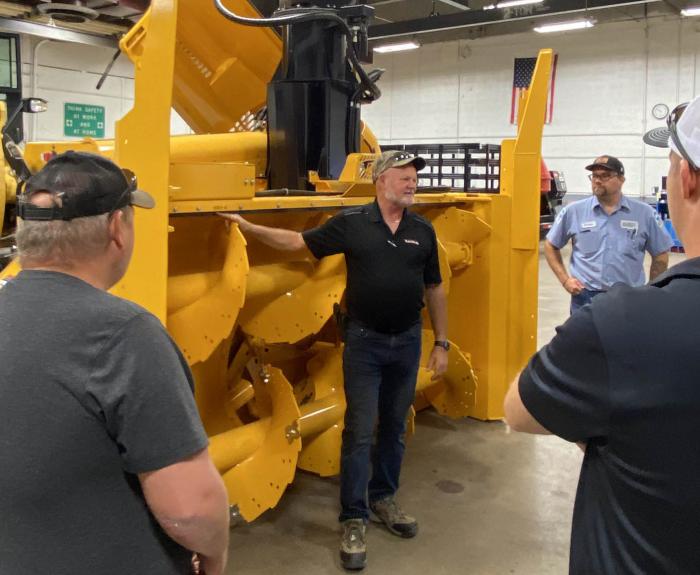 A group of people are gathered around a heavy-duty snowblower and receiving training on how to use the snowblower.