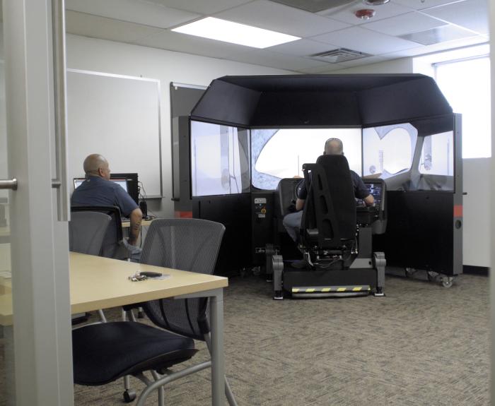 Man operating a snowplow simulator while another observes his progress