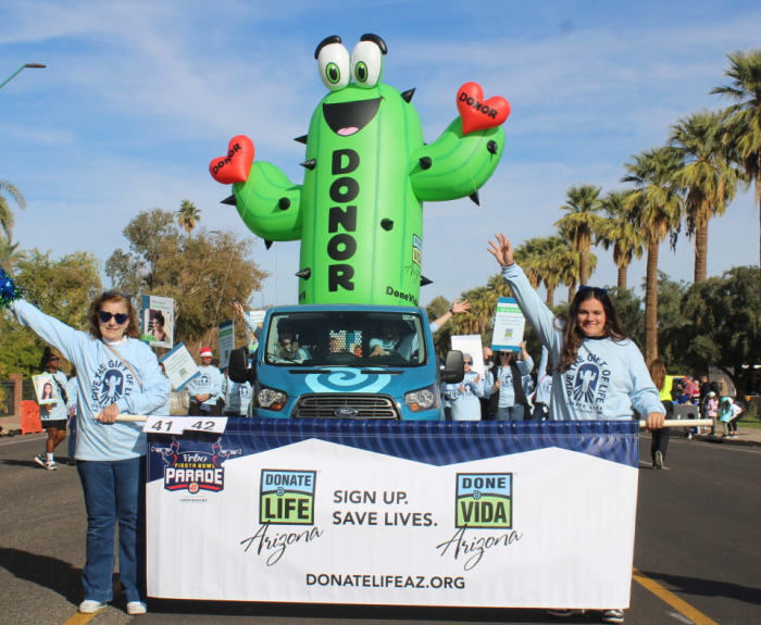 People walk in a parade down a city street. An very tall inflatable cactus can be seen.