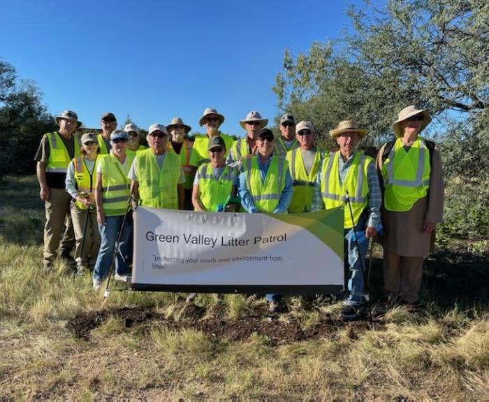 A group of people, all wearing bright yellow-green safety vests, gathers together outside and poses for a group photo during a volunteer clean-up effort along a highway.