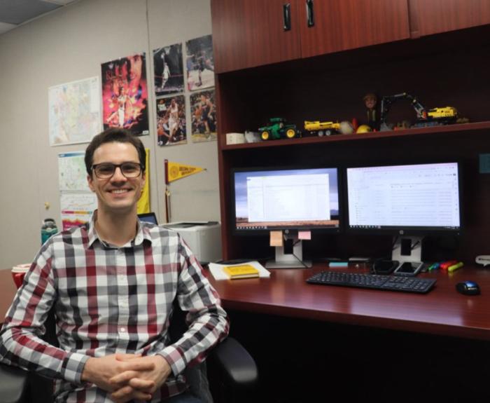 A man sits in a chair in front a desk with computer monitors in an office.
