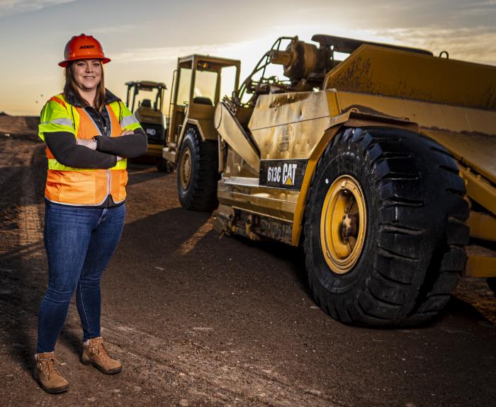 A woman in an orange reflective safety vest stands in a construction area with heavy equipment behind her.