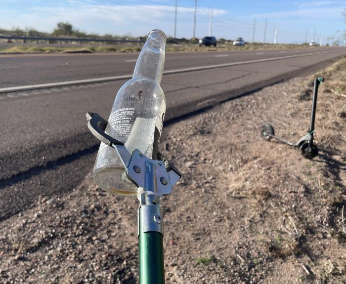 During a litter cleanup on the shoulder of a rural highway, an empty glass bottle is held by a litter grabber stick.