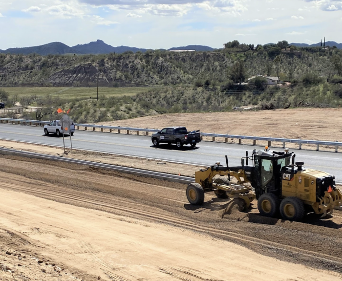 Heavy equipment works in a highway construction area as other vehicles drive by.