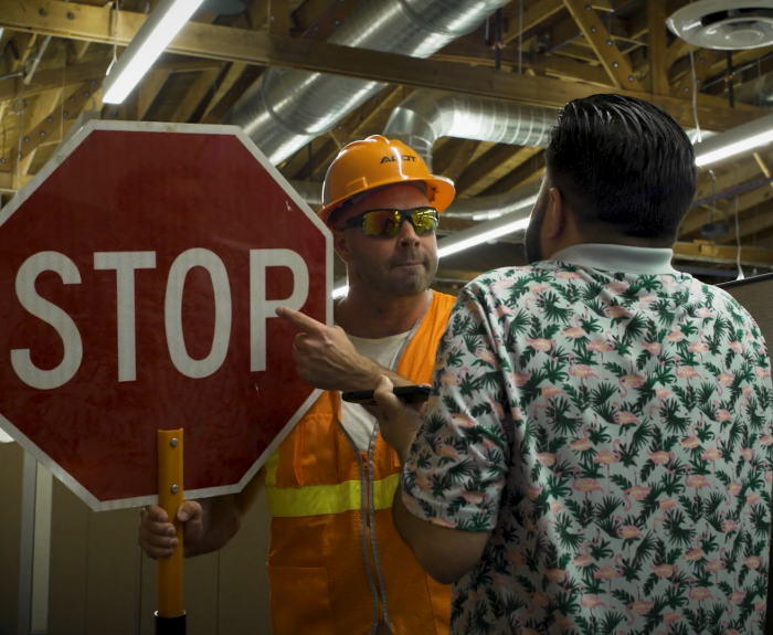 A man in a hard hat and orange vest points at a STOP sign while another man steps back.