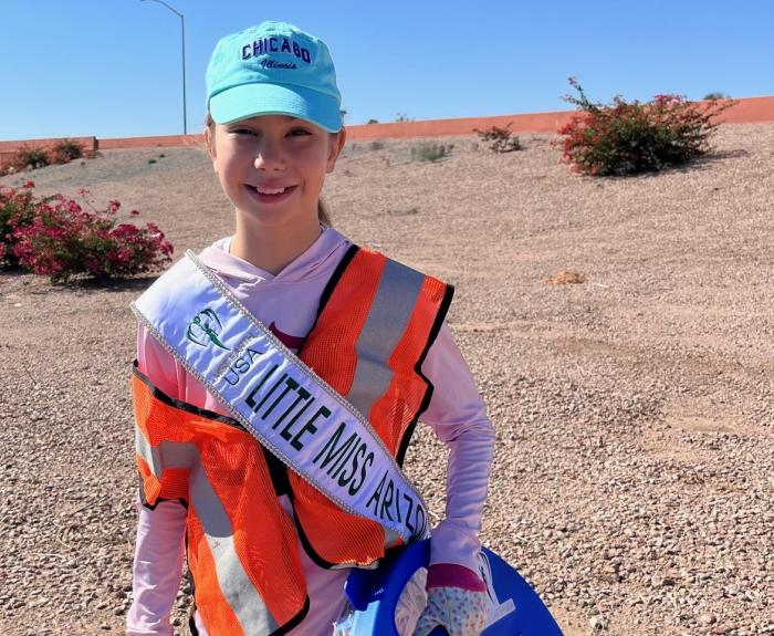 A girl wearing an orange reflective vest poses while picking up litter near a highway.