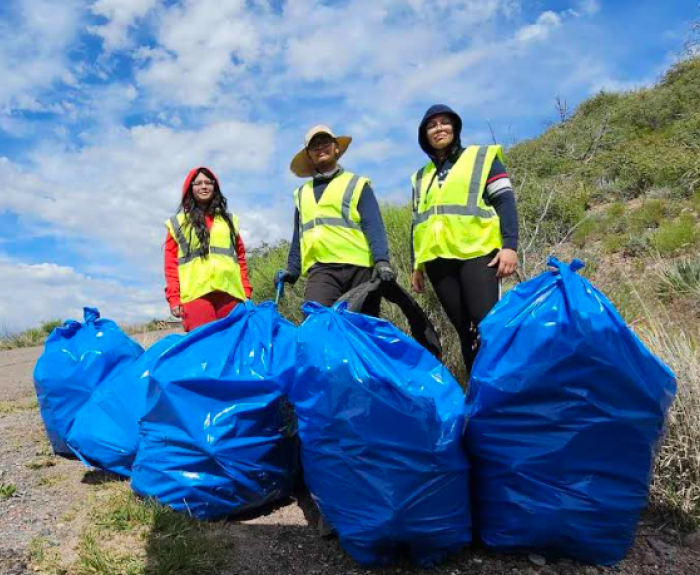 Three people stand behind large, blue trash bags filled with litter removed from a rural highway.