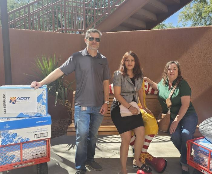 Three people pose for a photo in front of a bench and next to boxes filled with aluminum cab tabs.