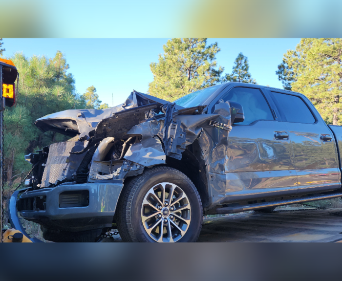 A black truck has severe front end damage. A blue sky and green trees are in the background.