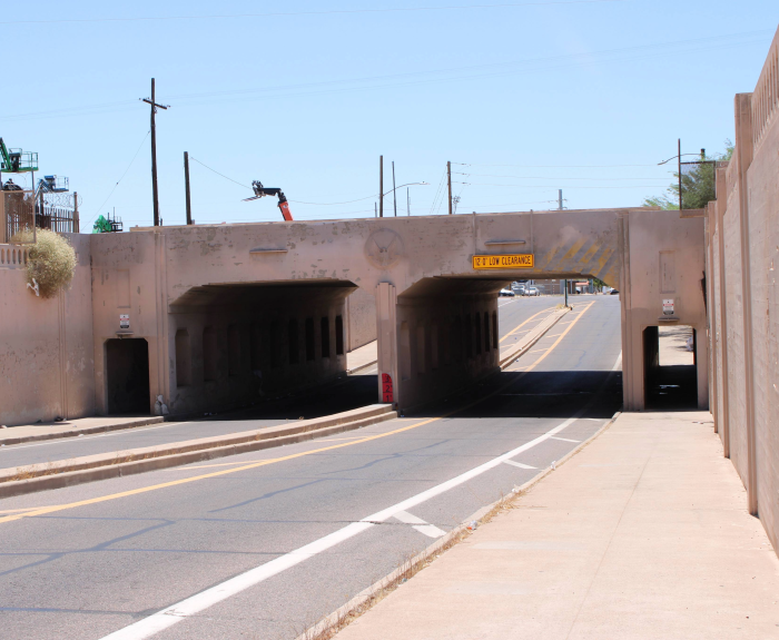 A roadway with two low-clearance underpasses, bordered by a sidewalk and a fence, stretches into the distance under a bright, clear sky.
