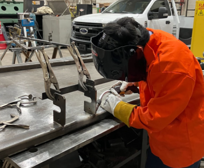 An individual in an orange safety jacket holding a welder and wearing a welding mask. 