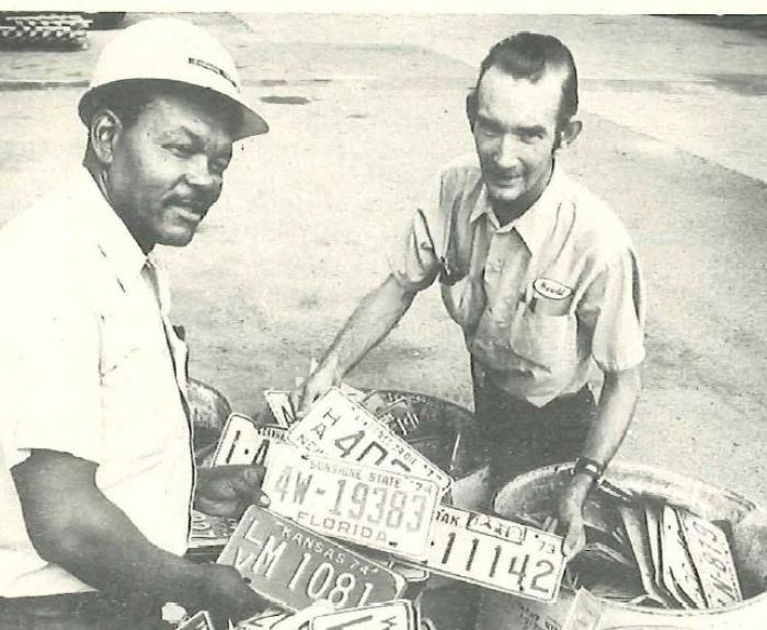 Two men stand next two barrels filled with license plates.