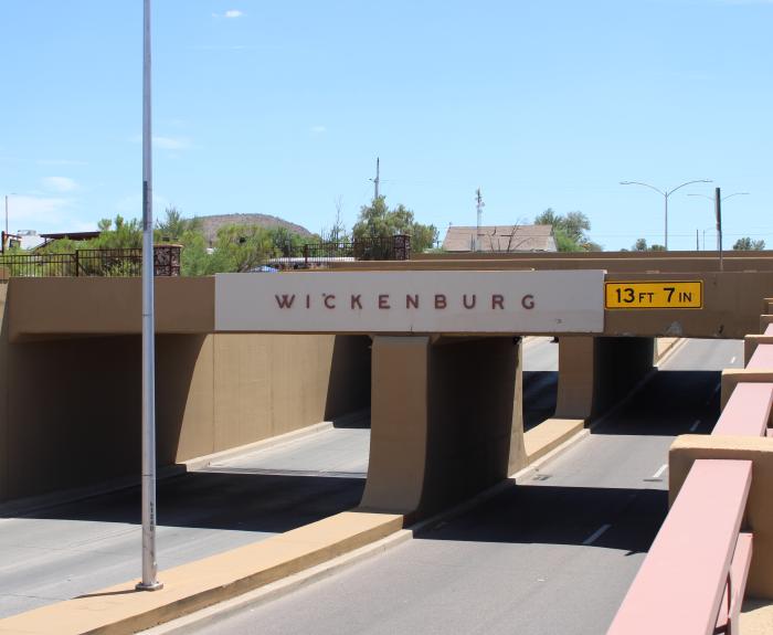 A road passes beneath a bridge with the name "Wickenburg" displayed on the side. A height restriction sign nearby indicates a clearance of 13 feet 7 inches.