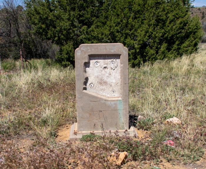 A stone marker along US 60 in Arizona is missing a metal plaque. The landscape is full of weeds, plants and a bush in the background.