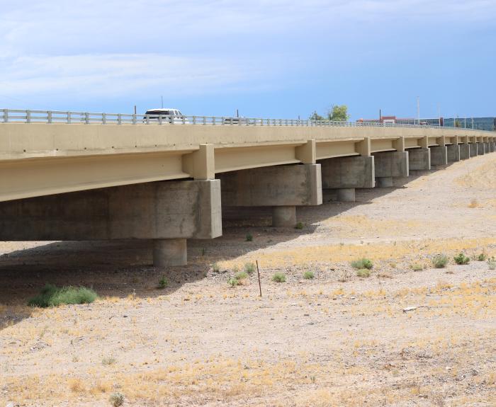 A concrete highway bridge spans a dry, sandy landscape under a cloudy sky. A vehicle is seen traveling on the bridge.