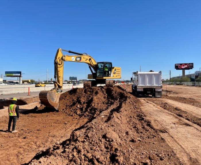 Construction site with an excavator moving dirt into a dump truck. A worker in a reflective vest stands nearby. Clear blue sky in the background.