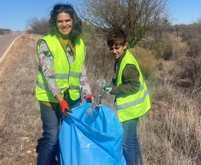 Two people on the shoulder of a rural two-lane highway collect litter and hold a big, blue garbage bag.