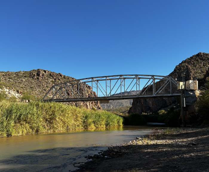 A steel truss bridge spans a calm river with rocky hills and clear blue sky in the background.