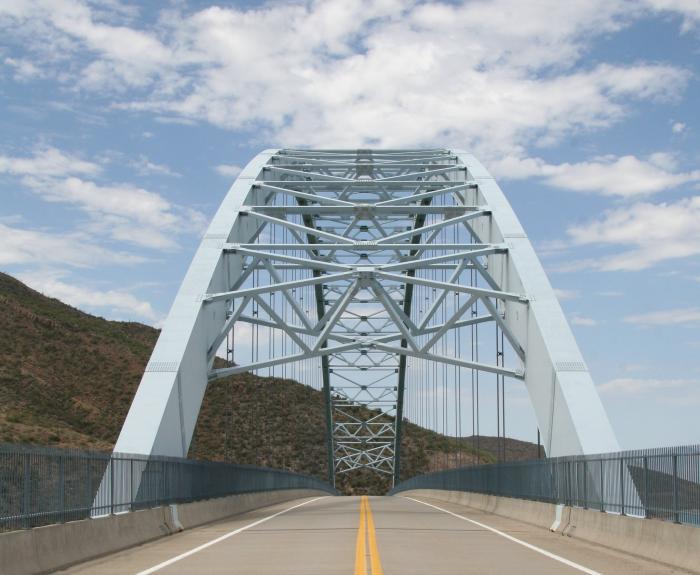 View of a steel arch bridge with a double yellow line road, surrounded by hills and a partly cloudy sky.