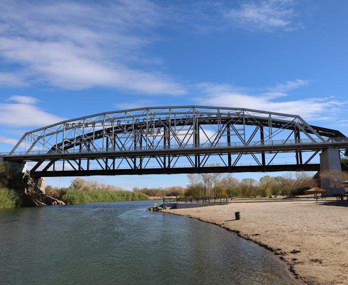 A steel truss bridge spans over a river with a sandy shore and shrubs. A clear blue sky with scattered clouds is in the background.