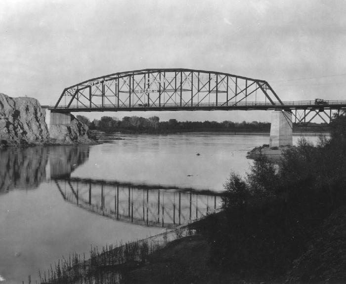 Black and white photo of a truss bridge spanning a calm river, with trees and rocky terrain on either side. The bridge's reflection is visible in the water.