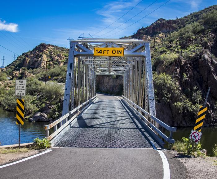 Metal truss bridge over a river, flanked by rocky hills, marked with a clearance sign of "14FT-3IN." Road signs and guardrails are present.