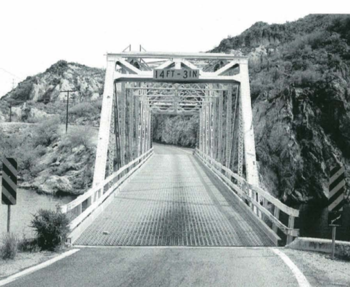 Metal truss bridge over a river, flanked by rocky hills, marked with a clearance sign of "14FT-3IN." Road signs and guardrails are present.