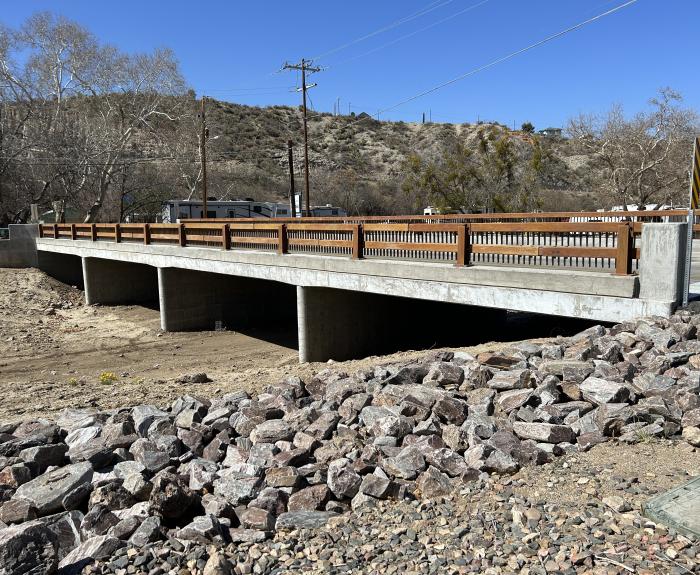 Concrete bridge with brown railings spans a dry, rocky creek bed. Power lines and barren trees are visible in the background under a clear blue sky.