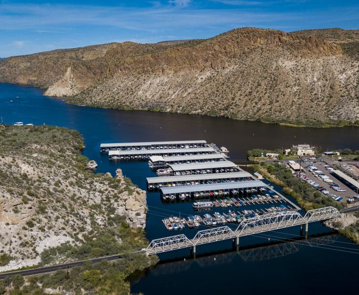 Aerial view of a marina with covered boat docks, a bridge, parking lot, and surrounding desert landscape by a river and rocky hills.