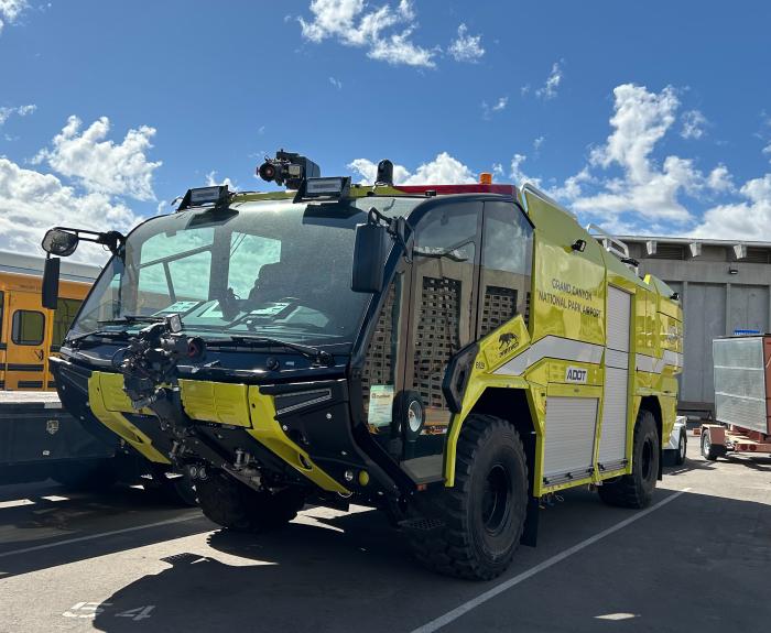 Large yellow airport fire truck labeled "ADOT" parked outdoors on asphalt under a partly cloudy sky, with industrial buildings in the background.