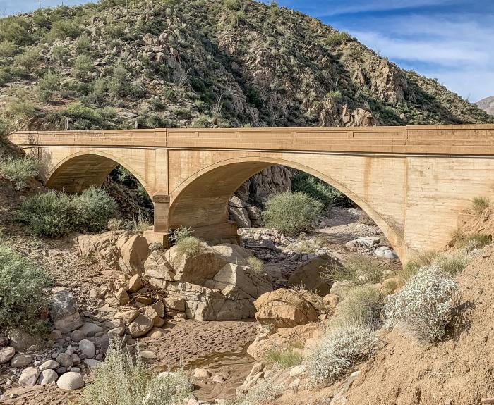 A concrete arch bridge spans a dry, rocky creek bed surrounded by desert vegetation and hills.