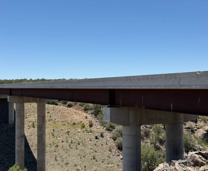 A concrete bridge with large support pillars spans over a dry, rocky landscape under a clear blue sky.