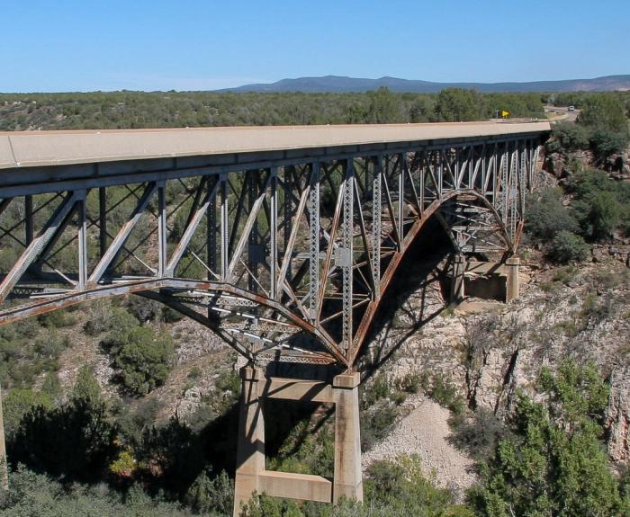 A steel truss bridge spans a rocky canyon with sparse vegetation and distant hills under a clear blue sky.