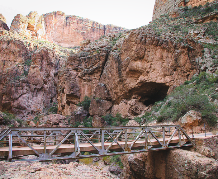 A metal footbridge spans a rocky canyon with steep cliffs and sparse vegetation under a bright sky.