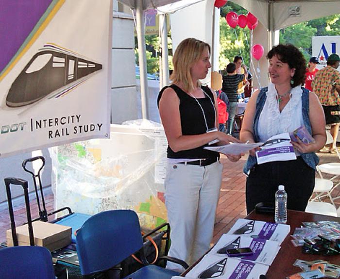 Two women stand at a booth with pamphlets and a banner reading ADOT Intercity Rail Study at an outdoor event.