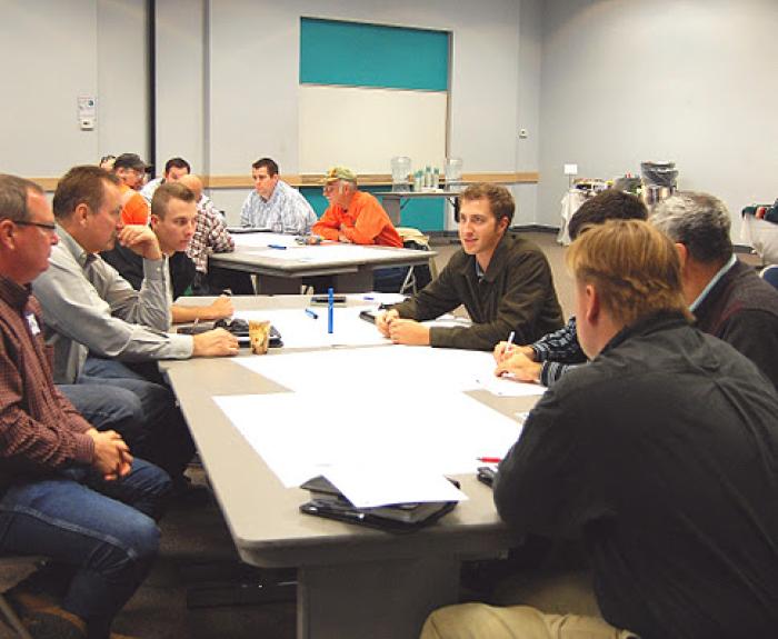 A group of people sit around tables in a meeting room, engaged in discussion and writing on paper.