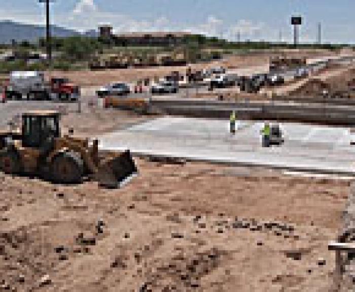 Construction workers pour and smooth concrete on a roadway project, with heavy machinery and vehicles nearby under clear skies.