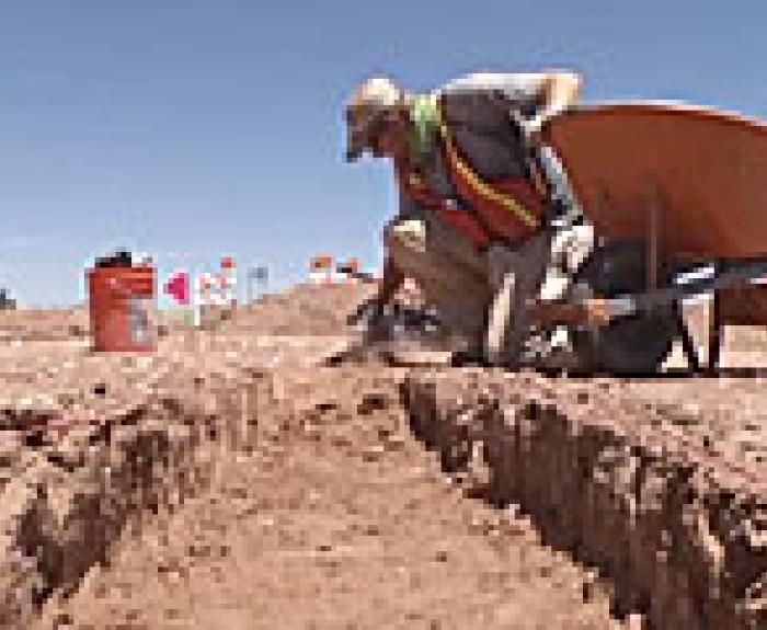 Person wearing safety gear excavates a trench at an archaeological site under a clear sky, with a wheelbarrow and orange bucket nearby.