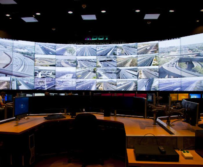 A control room with large display screens showing multiple live surveillance camera feeds of highways and roads, surrounded by workstations and computer monitors.