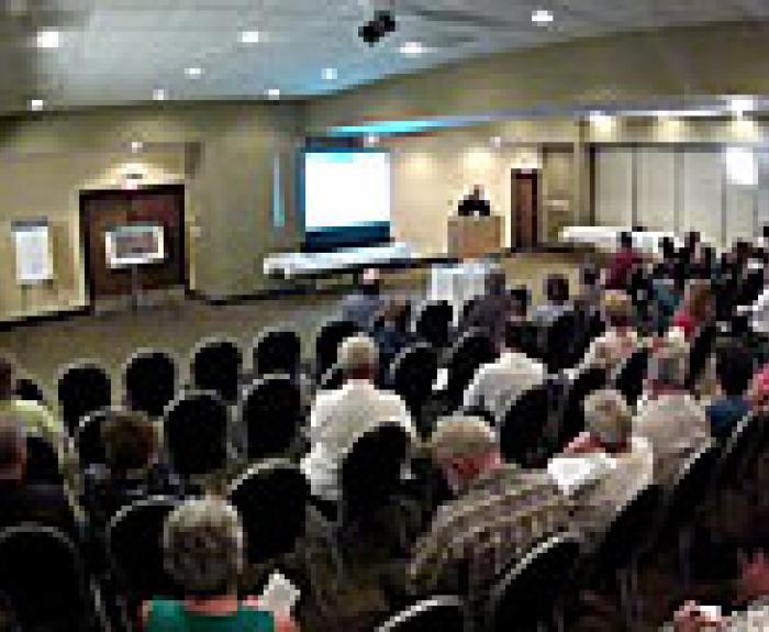 A speaker presents to an audience seated in a conference room with a projection screen at the front.
