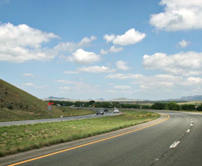 A curved highway with cars driving under a blue sky with scattered clouds and grassy hillsides.