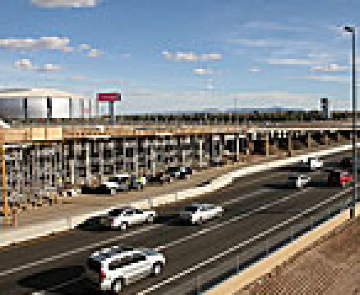 Cars driving on a highway next to an overpass under construction, with a stadium in the background.