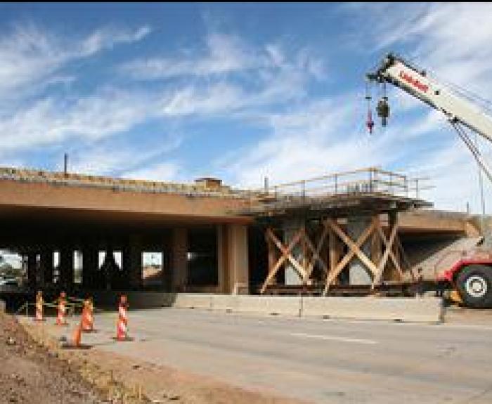 A crane and construction scaffolding are positioned near an overpass under repair, with traffic cones blocking part of the road.