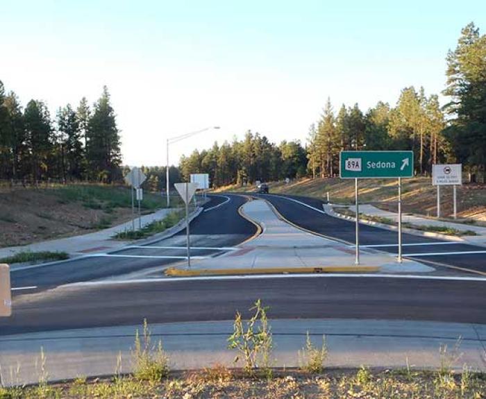 A newly paved road with a roundabout, surrounded by trees. A green highway sign indicates the direction to Sedona via State Route 89A.