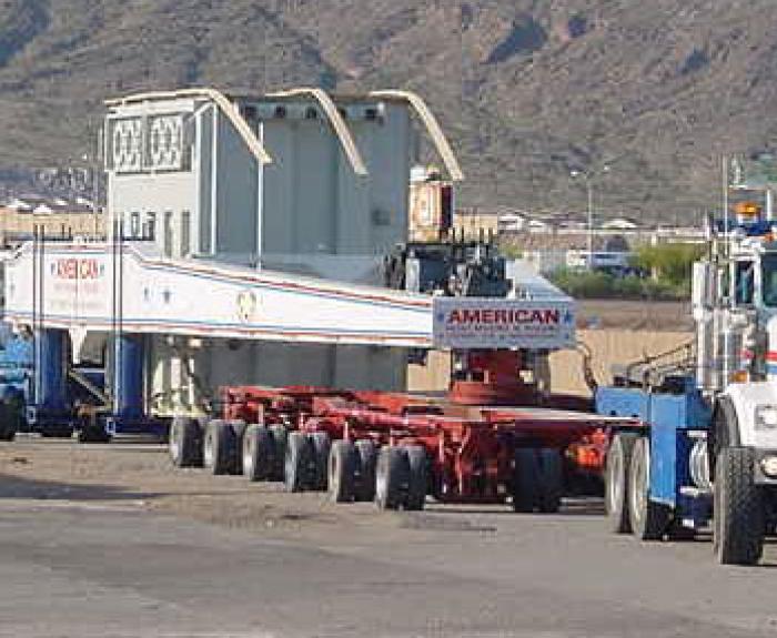 Three large trucks transport an oversized load on a multi-axle trailer along a road near mountains.