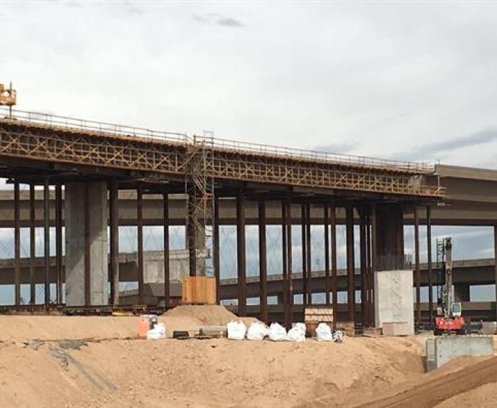 A highway overpass under construction with scaffolding, cranes, and sand piles in the foreground.