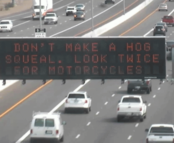 Overhead highway sign reads, Dont make a hog squeal. Look twice for motorcycles, above lanes of moving traffic.