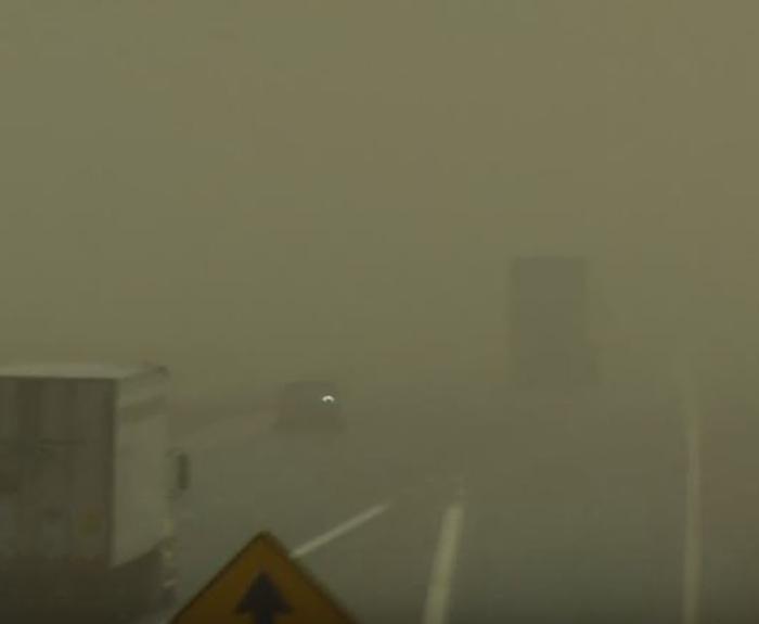 Trucks and vehicles drive on a highway with very low visibility due to heavy dust making road signs and surroundings barely visible.