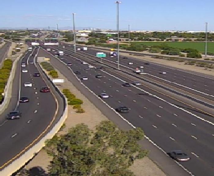 A wide freeway with moderate traffic flows beside fields and trees under a clear blue sky.