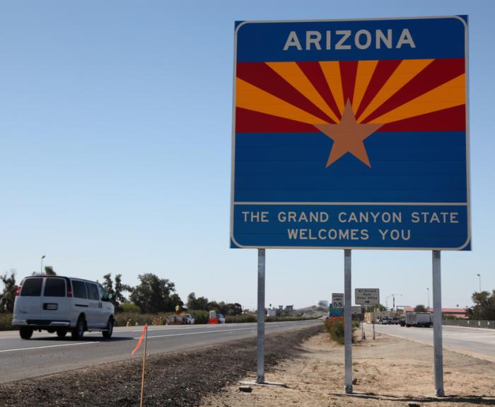 A blue Arizona welcome sign by a highway reads The Grand Canyon State Welcomes You.